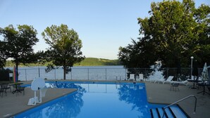 Una piscina con aro de baloncesto, sillas de descanso y vistas a un lago.