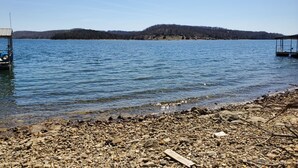 Vista de un lago con orilla rocosa, un muelle y una colina boscosa al fondo.
