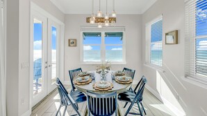A dining room with a round table set for six, a chandelier, and a view of the ocean through large windows.