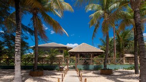 A wooden pathway leading to a building with a tiled roof, surrounded by palm trees and a clear blue sky. - VG Sun Cumbuco Pé Na Areia Bangalô 6 Pessoas By DM Apartments (Caucaia)