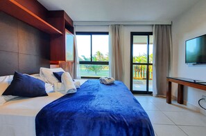 A hotel room with a large bed, a TV, and a balcony with a view of palm trees. - VG Sun Cumbuco Pé Na Areia Bangalô 6 Pessoas By DM Apartments (Caucaia)