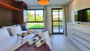 A hotel room with a bed, a TV, a balcony with a view of a golf course, and towels with a logo. - VG Sun Cumbuco Pé Na Areia Bangalô 6 Pessoas By DM Apartments (Caucaia)