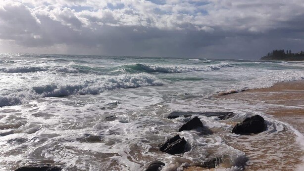 Una playa con olas rompiendo en la orilla, cielo despejado y una línea de costa distante con edificios.