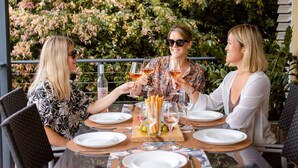 Tres personas sentadas en una mesa al aire libre con copas de vino, platos y una tabla de madera con alimentos.