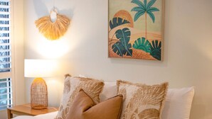 Una cama bien hecha con una colcha blanca y dos almohadones decorativos, una obra de arte en la pared que representa follaje tropical, y una lámpara de pared.