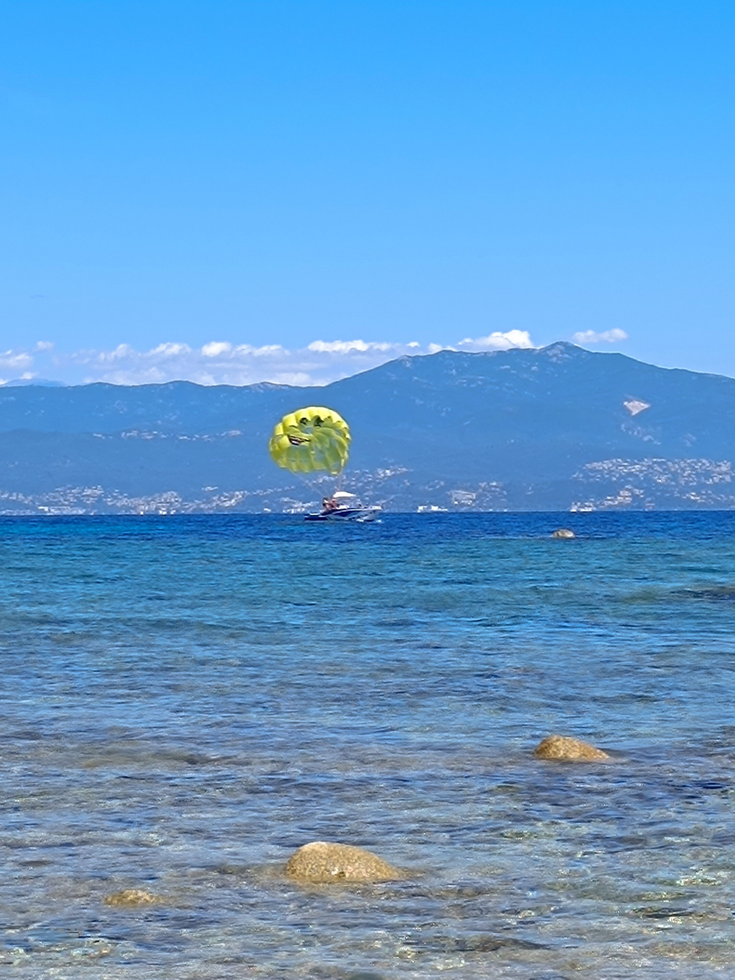 On peut clairement voir une plage aux eaux calmes, un parachute ascensionnel dans le ciel et une chaîne de montagnes en arrière-plan.