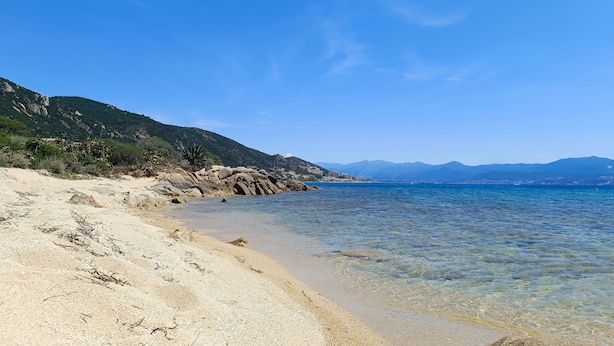 Ein Strand mit klarem Wasser, felsigem Ufer und Bergen im Hintergrund.