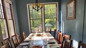 Una sala menjador amb una taula llarga preparada per a una menjada, grans finestres amb vista a vegetació, i un penjoll.