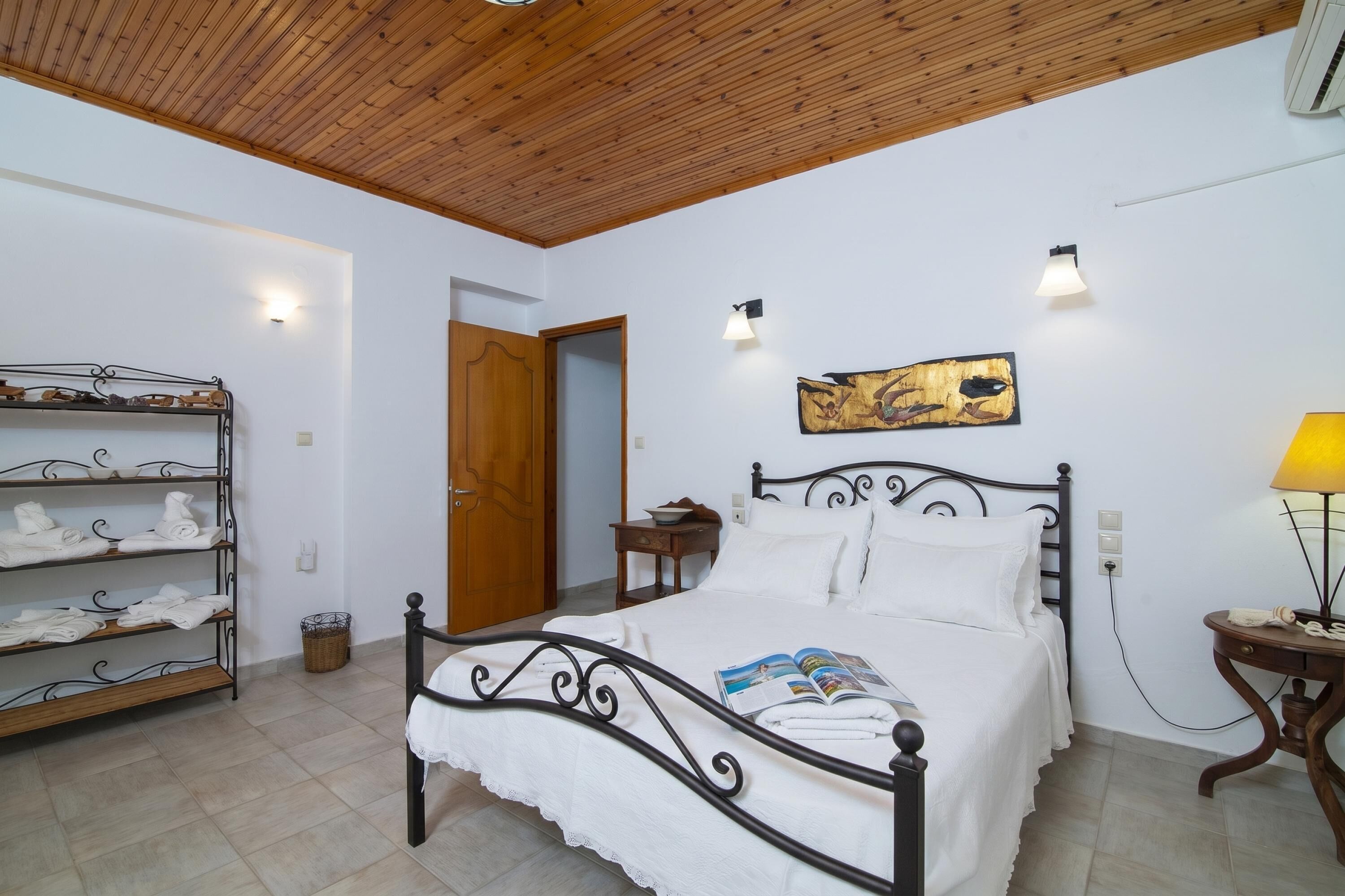 Une chambre à coucher avec un lit, une étagère où sont pliées des serviettes, une table de chevet avec une lampe et un tableau accroché au mur.