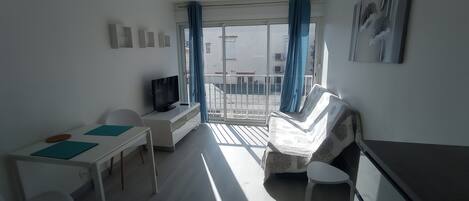 A modern, well-lit room with a sofa, a TV, a desk, and a balcony with blue curtains.