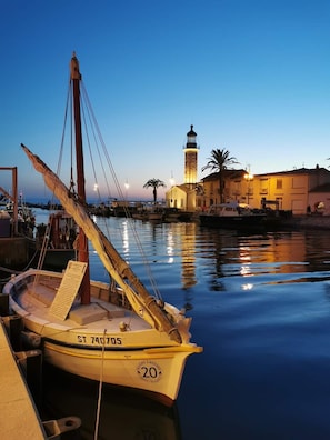 Un port tranquille au crépuscule, avec un bateau amarré au quai, un phare en arrière-plan et un bâtiment doté d’un balcon donnant sur l’eau.