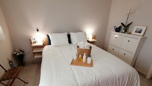 En pænt redt seng med hvide sengetøj, et natbord med en lampe, en lille plante og et indrammet billede på sengebordet.