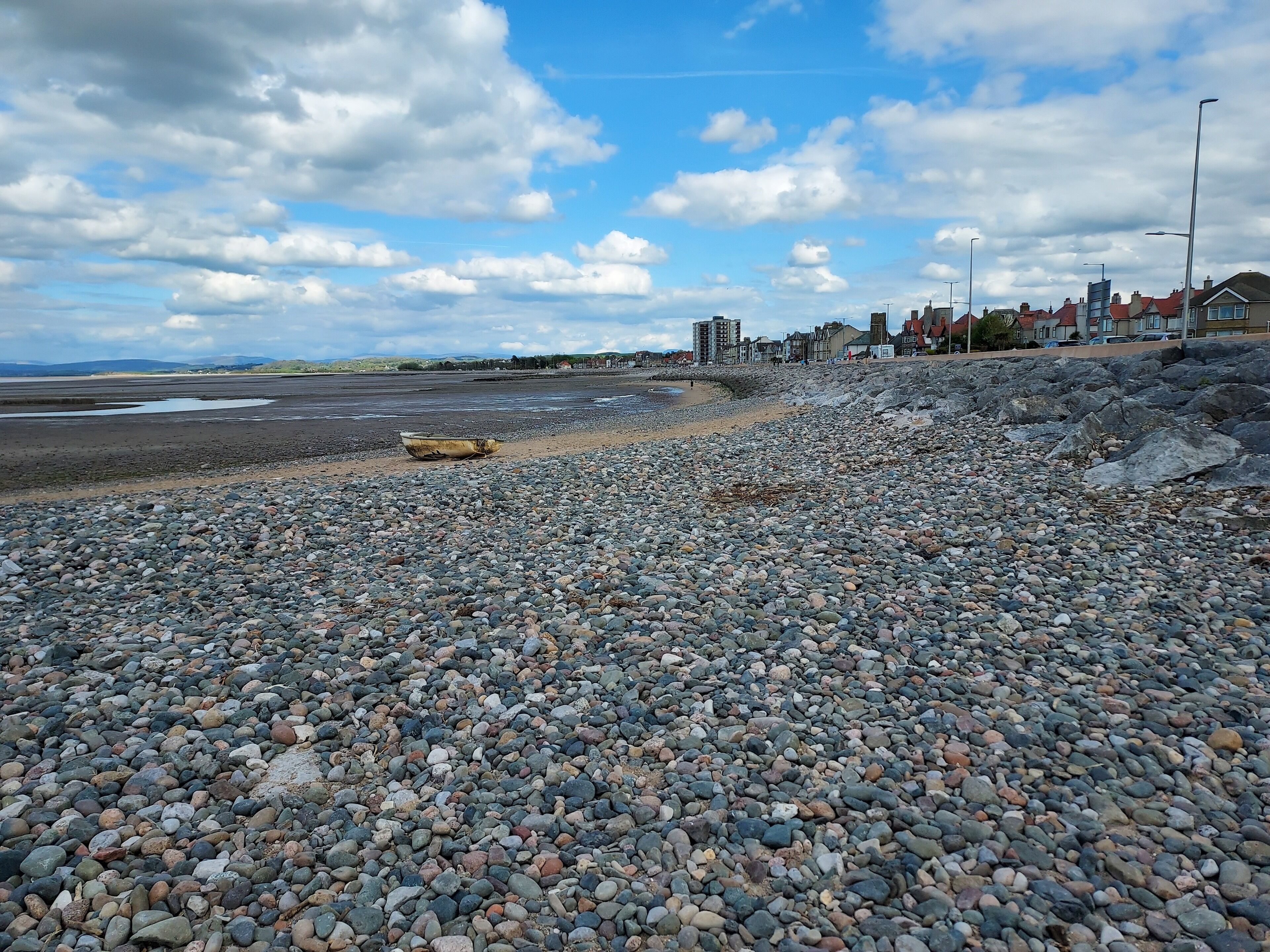A pebbled beach with a boat, rocky shore, and buildings in the background.
