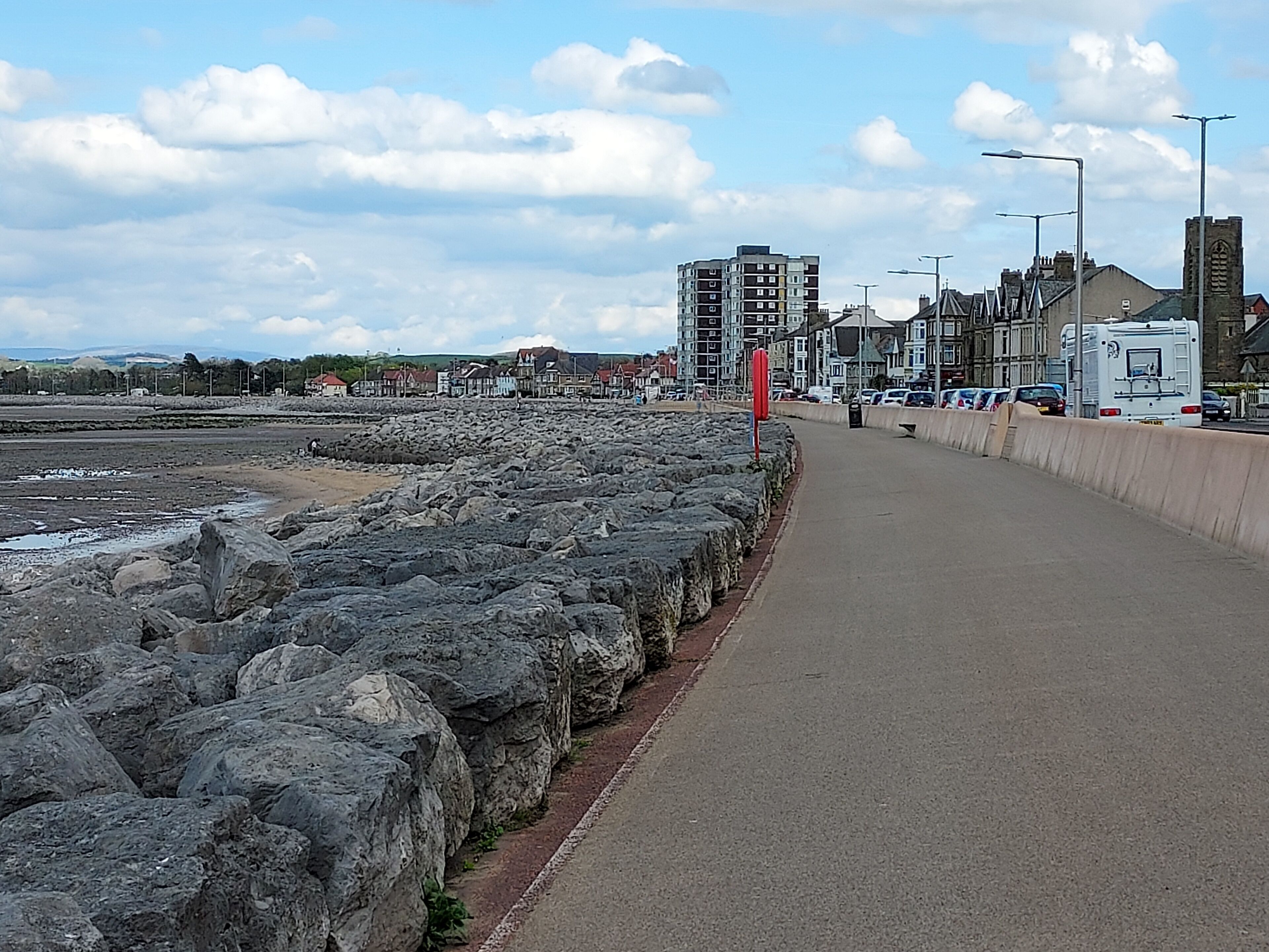 Un passeig marítim amb un mur de pedra a la vora del mar, un camí pavimentat i edificis al fons.