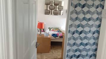 Une pièce comprenant un lit, une commode en bois, une lampe et un mur orné d’un motif géométrique.