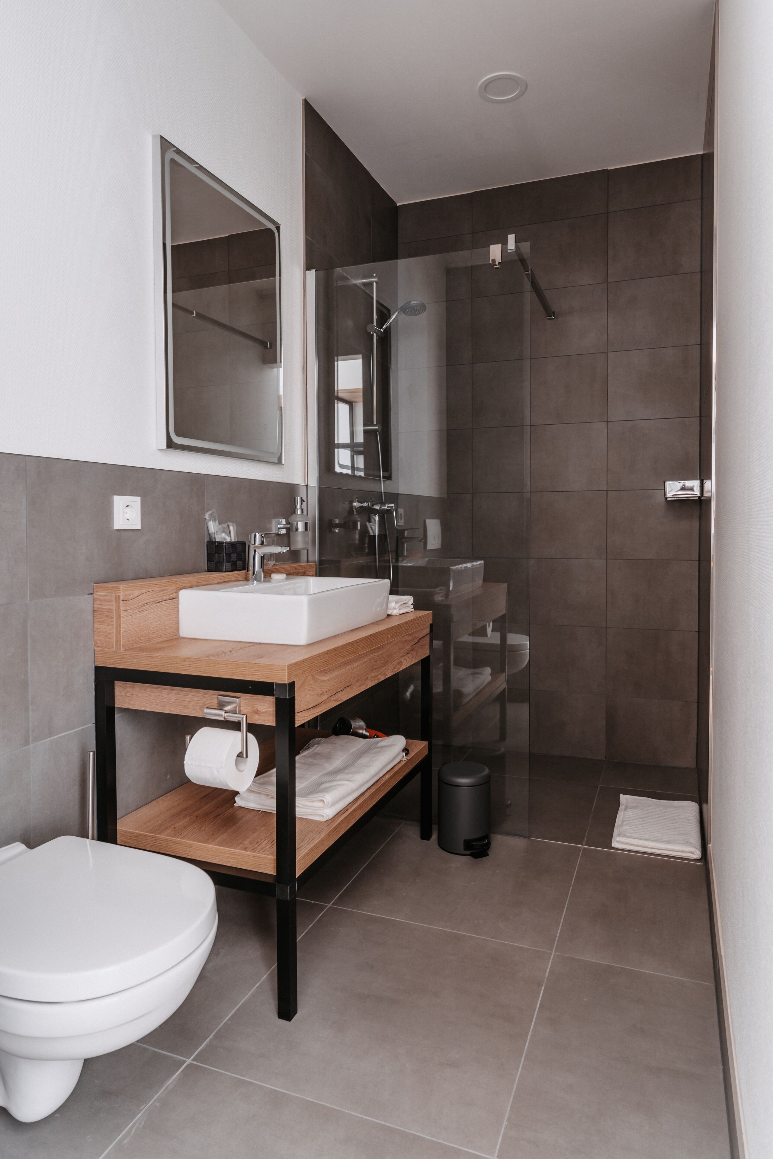 A modern bathroom with a wooden vanity, a white sink, a mirror, a toilet, and a glass-enclosed shower area.