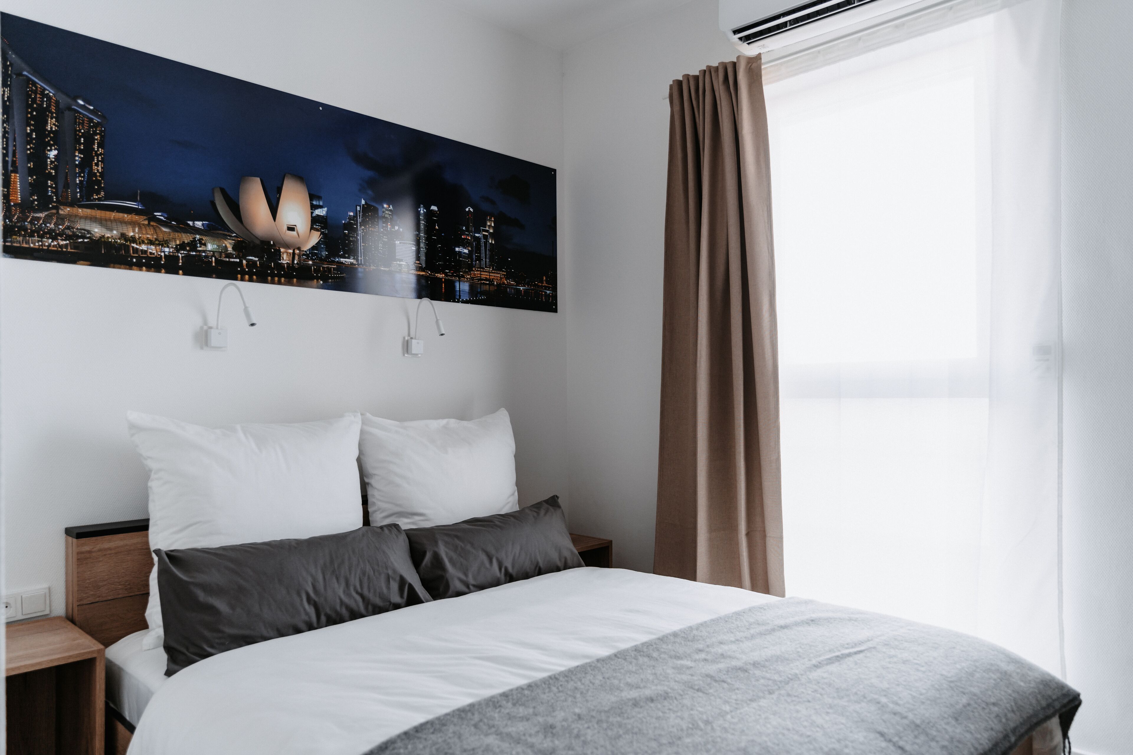 A hotel room with a bed, a nightstand, a wall-mounted lamp, a window with curtains, and a cityscape painting.