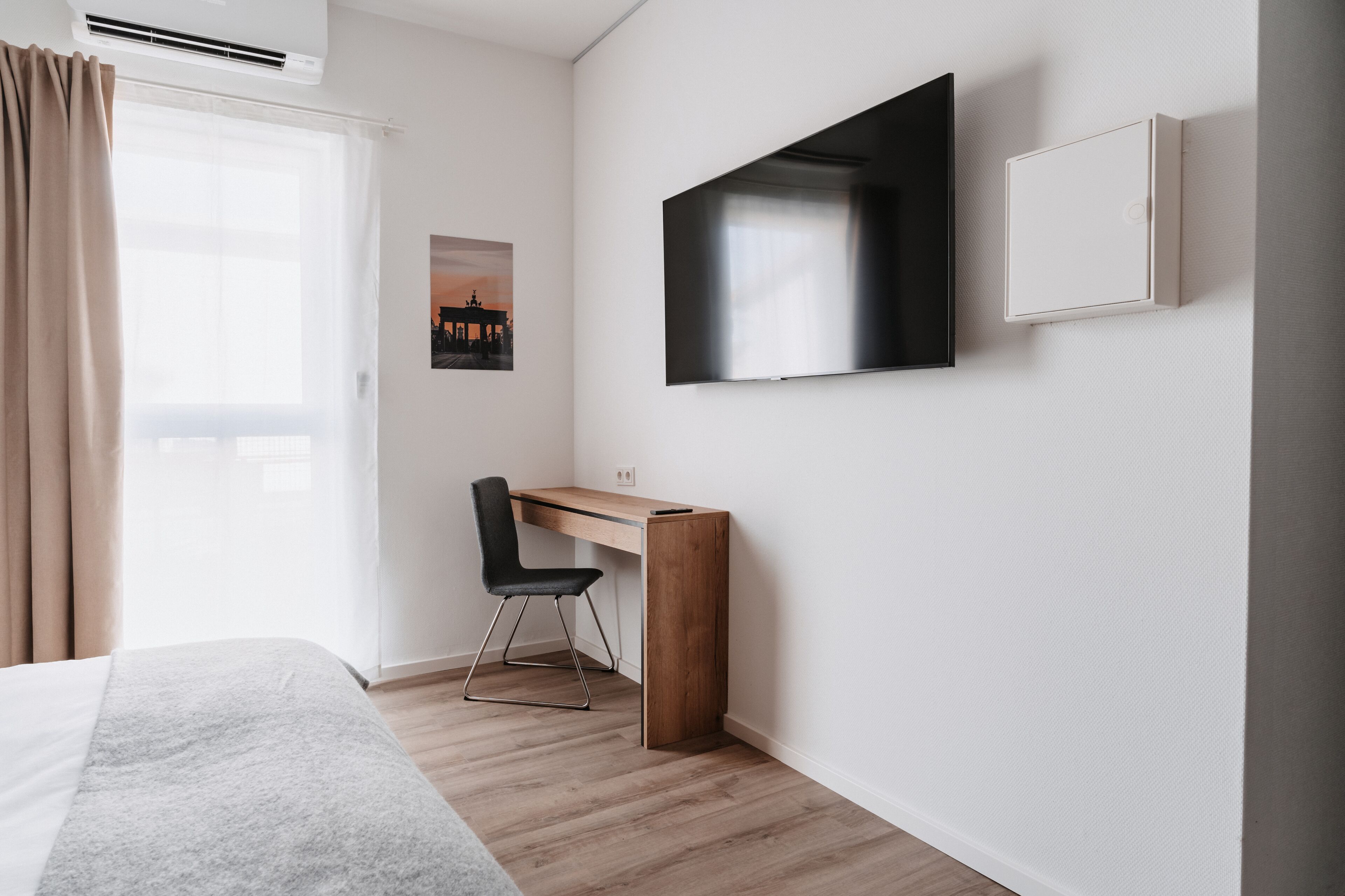 A modern hotel room with a bed, a desk with a chair, a flat-screen TV, and a window with curtains.