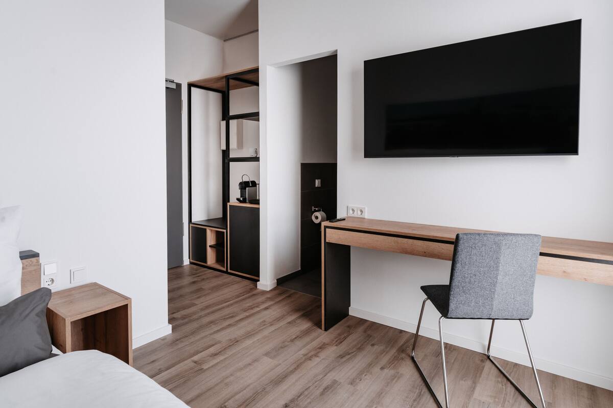 A modern hotel room with a wooden desk, a grey chair, a bed, and a TV mounted on the wall.