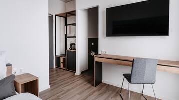 A modern hotel room with a wooden desk, a grey chair, a bed, and a TV mounted on the wall.