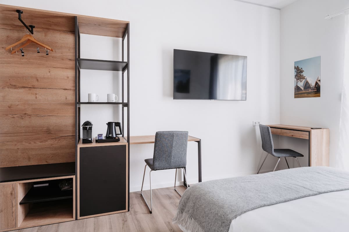 A modern bedroom with a bed, a desk, and a chair. There is a wardrobe with shelves, a hanging rack, and a coffee maker.