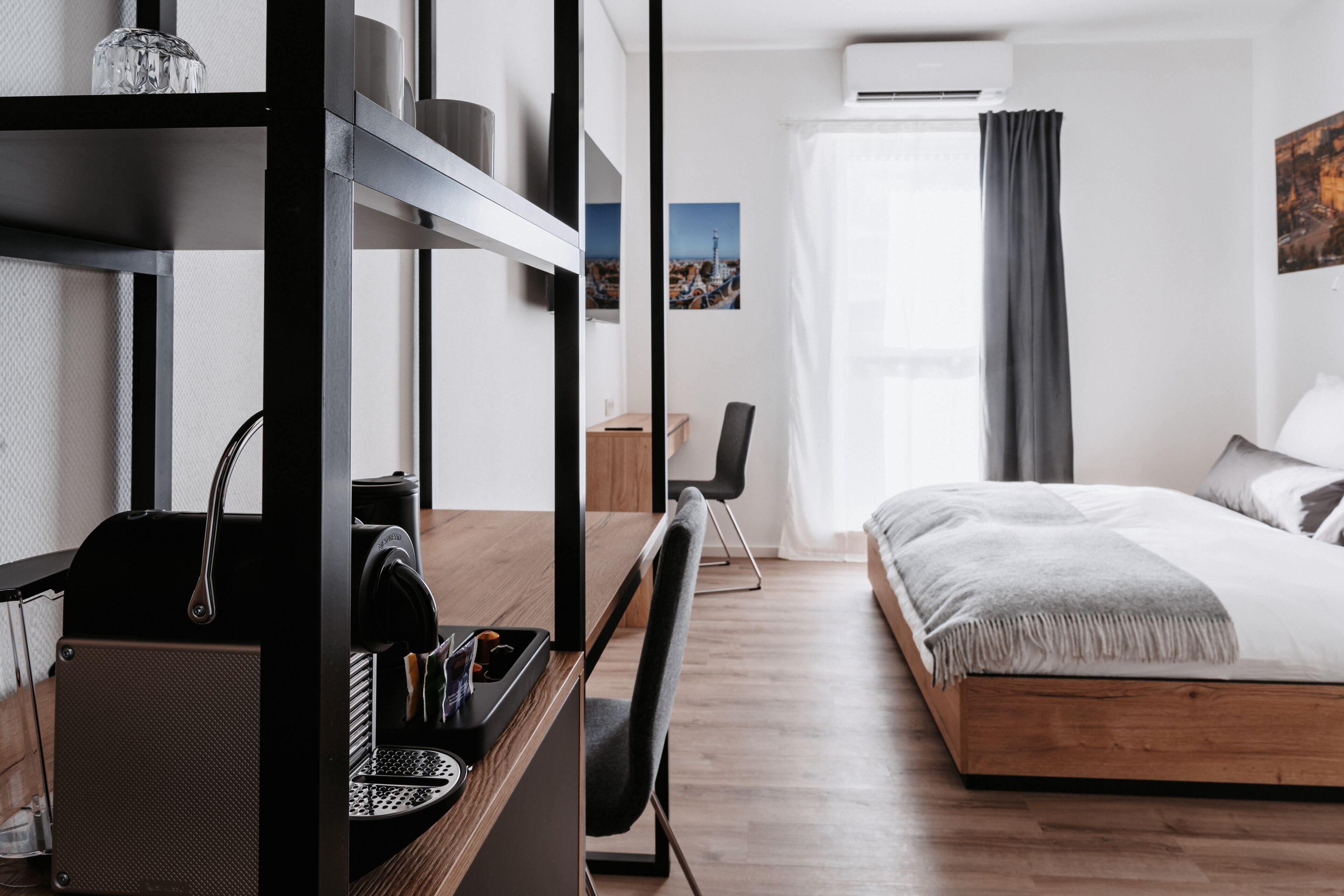 A modern hotel room with a wooden bed, a desk with a coffee machine, a chair, and a view of the city through large windows.