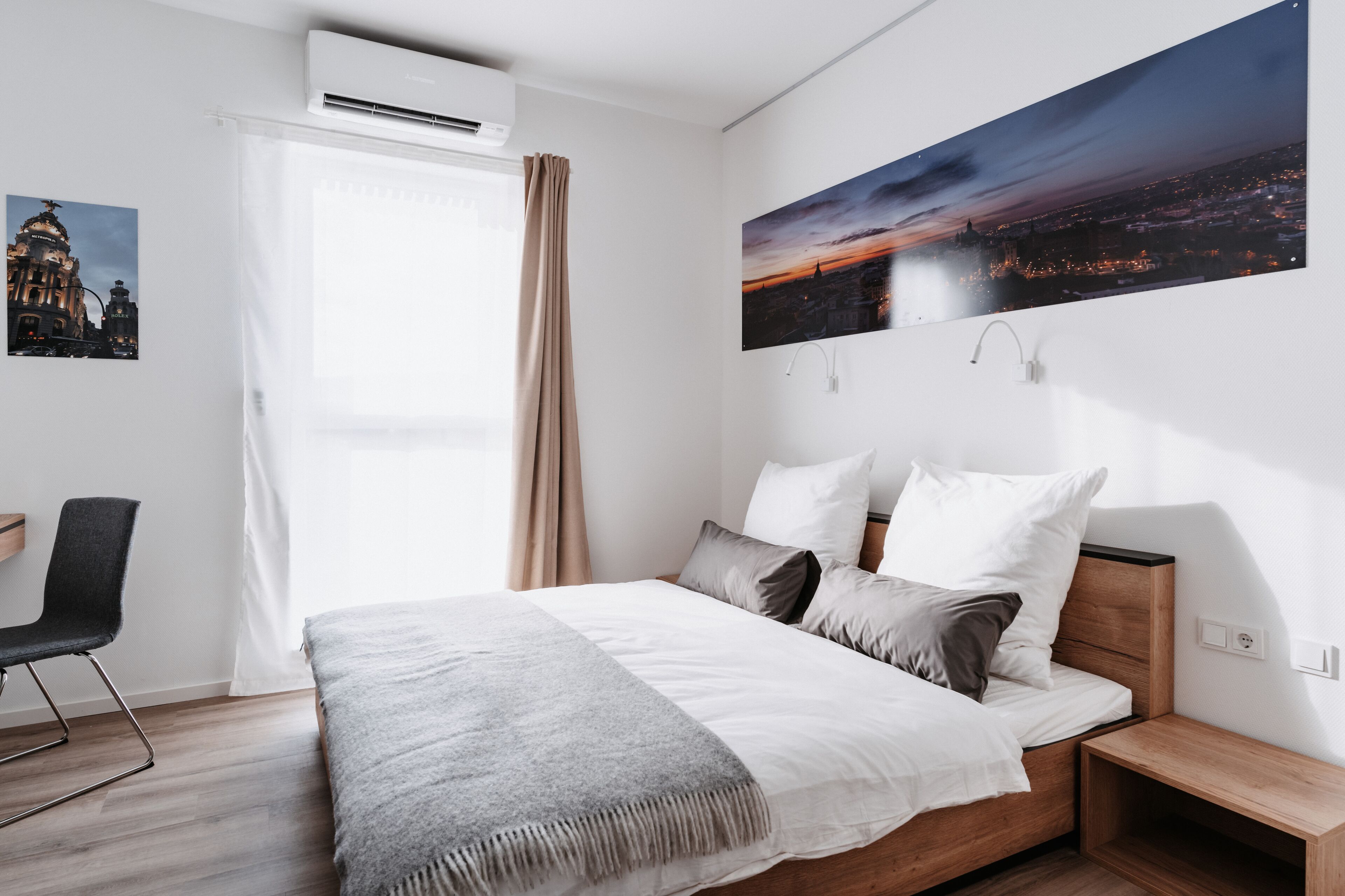 A modern bedroom with a large bed, a desk, and a cityscape view.