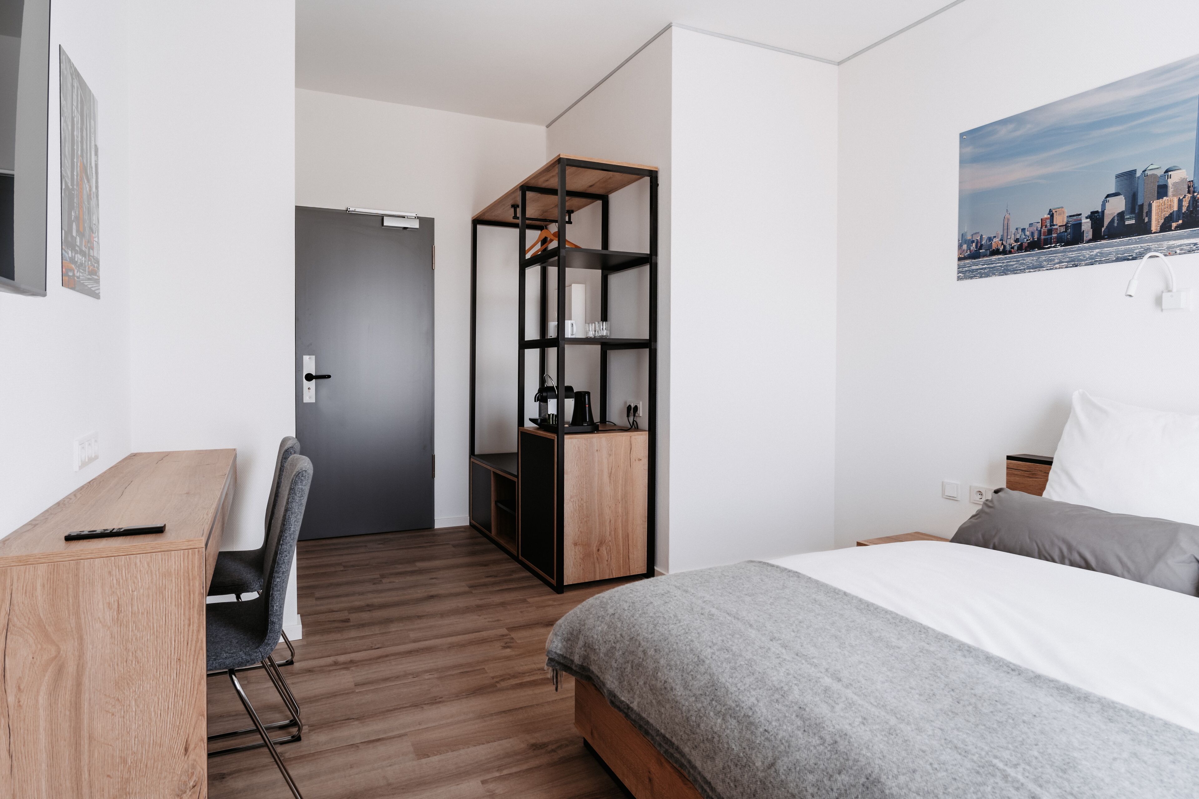 A modern hotel room with a wooden desk, a bed with grey bedding, a wardrobe with shelves, and a cityscape painting on the wall.