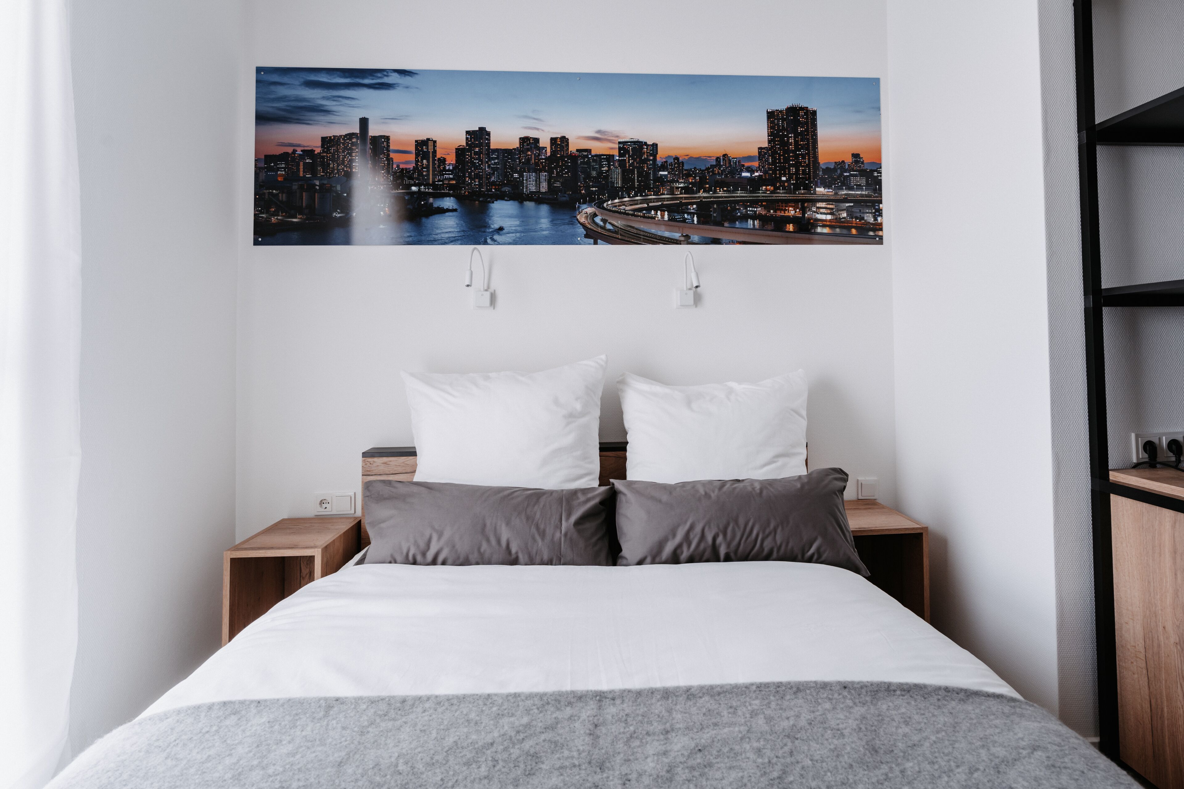 Een opgemaakt bed met wit en grijs beddengoed, twee nachtkastjes met lampen, en een stadsgezicht schilderij erboven.