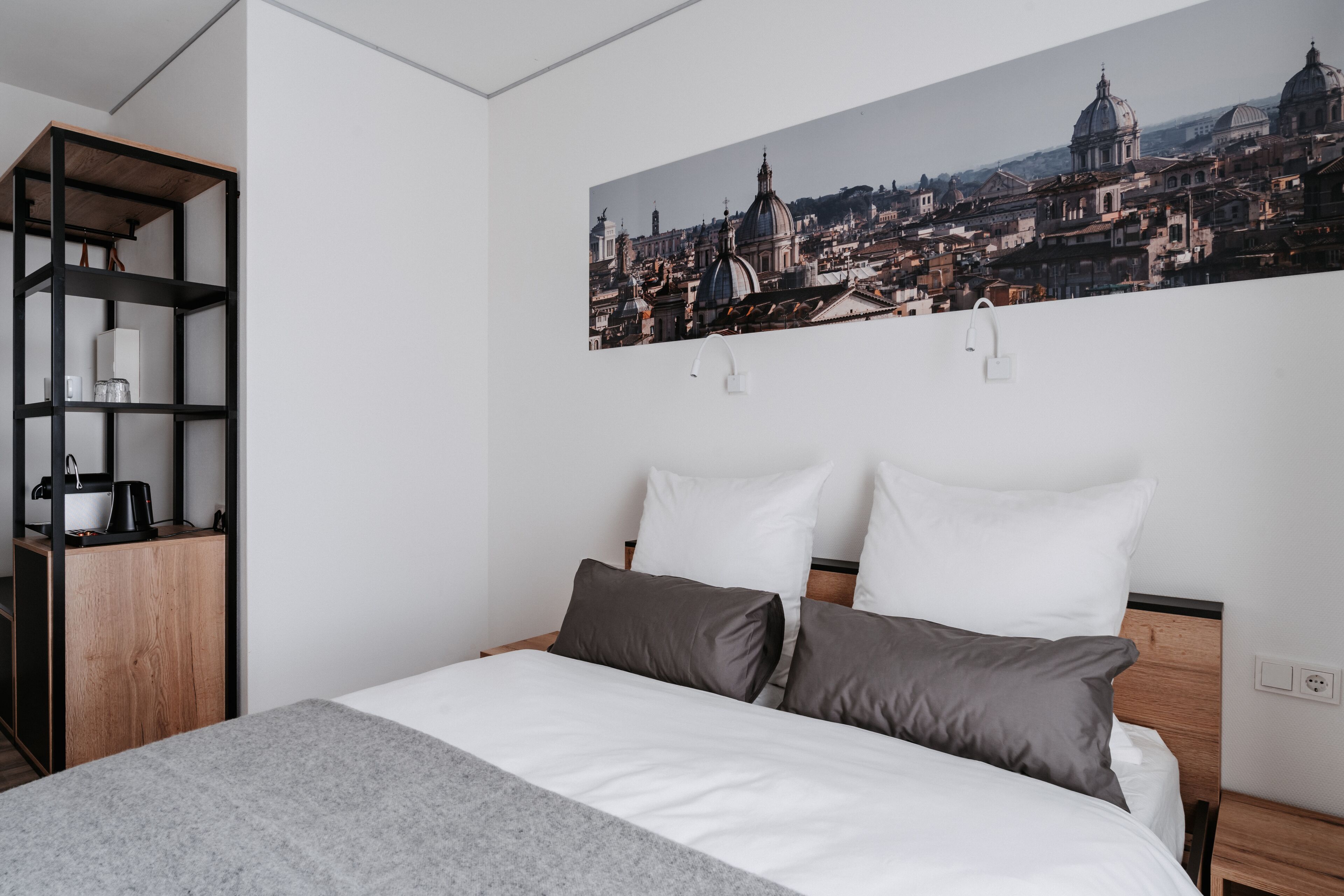 Habitación de hotel con cama, mesita de noche, estantería y cuadro de arte con vista urbana.