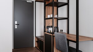 A compact room with a dark door, a wooden desk with a chair, and a built-in shelving unit with various items. - Step Inn The Fair Hotel (Rosbach vor der Hoehe)
