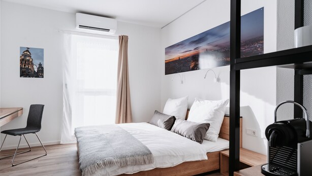 A modern hotel room with a wooden bed, a desk, and a chair. There are two paintings on the wall, a window with curtains, and an air conditioning unit. - Step Inn The Fair Hotel (Rosbach vor der Hoehe)