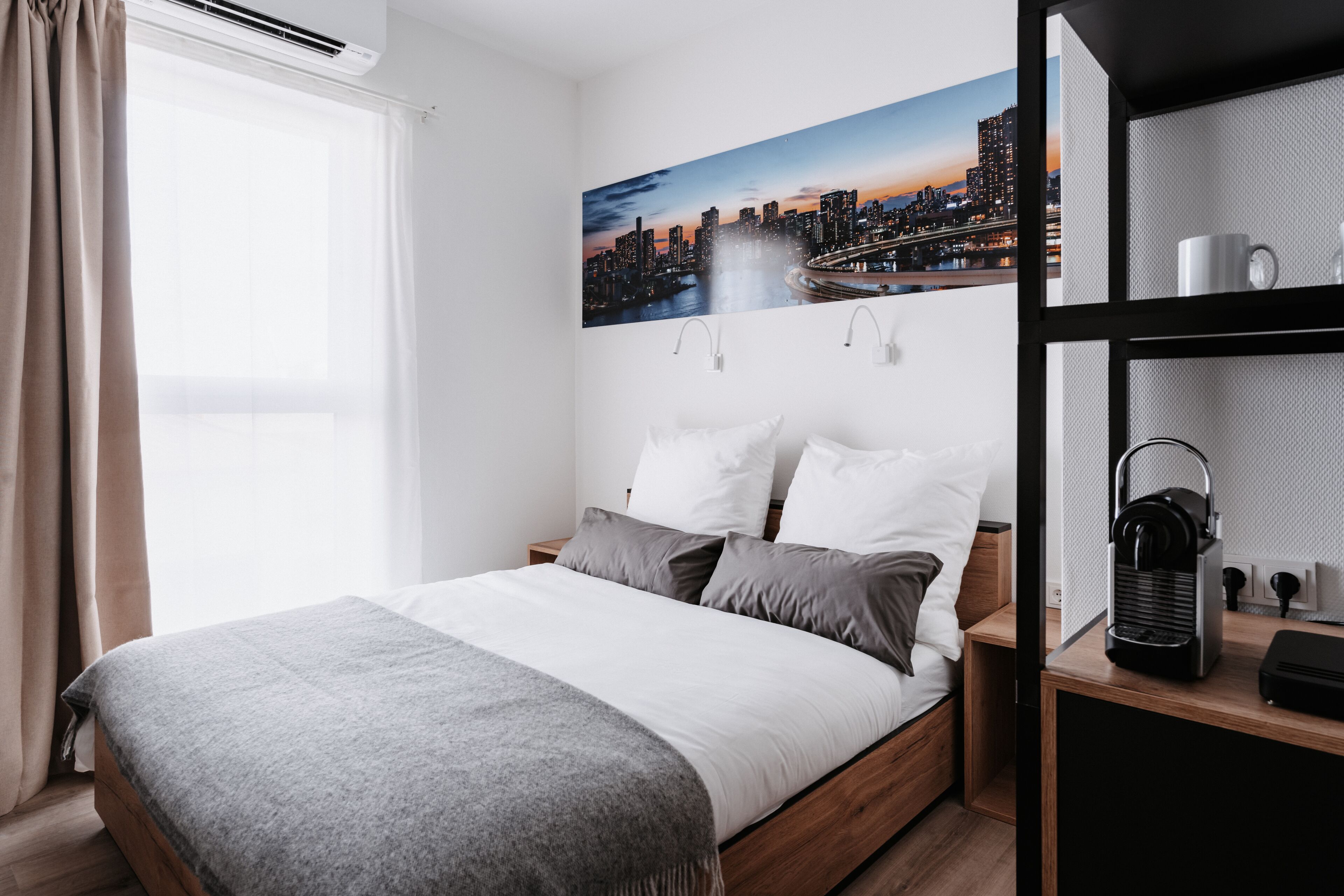 Une chambre d’hôtel avec un lit, un tableau représentant un paysage urbain, une table de chevet, une lampe et une étagère avec une bouilloire.