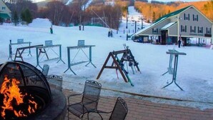 A snow-covered outdoor area with a fire pit, tables, and chairs, surrounded by buildings and a ski lift in the background. - Chic 2BR Suite on Jiminy Mtn Sleeps 8 - Gorgeous (Hancock)