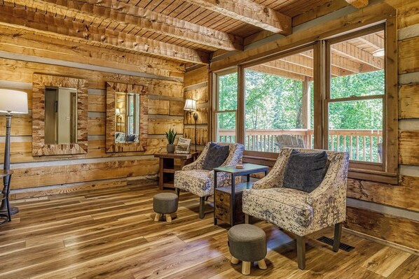 A cozy cabin interior with wooden walls, large windows, and a seating area with patterned chairs and ottomans.