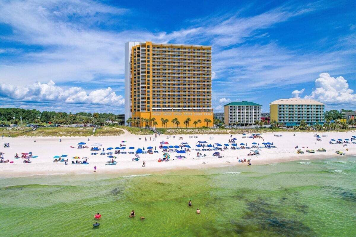 Un hotel frente a la playa con un edificio grande, una playa de arena con sombrillas y palmeras.