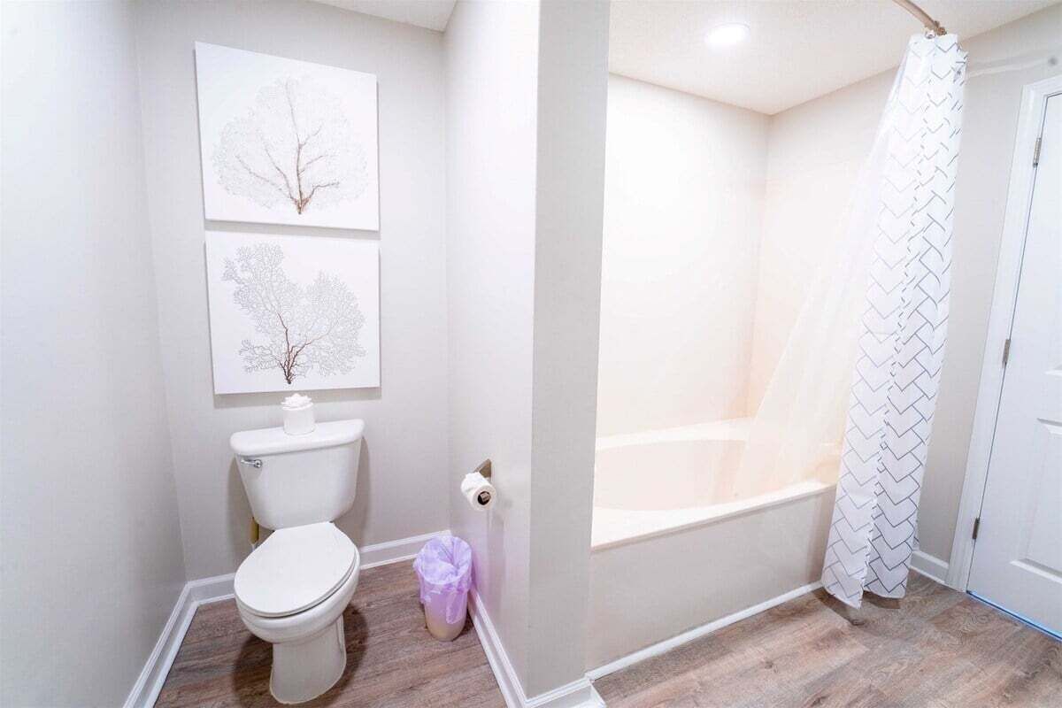 Une salle de bain comprenant des toilettes, deux œuvres d’art accrochées au mur, une poubelle et un espace douche avec un rideau à motifs.
