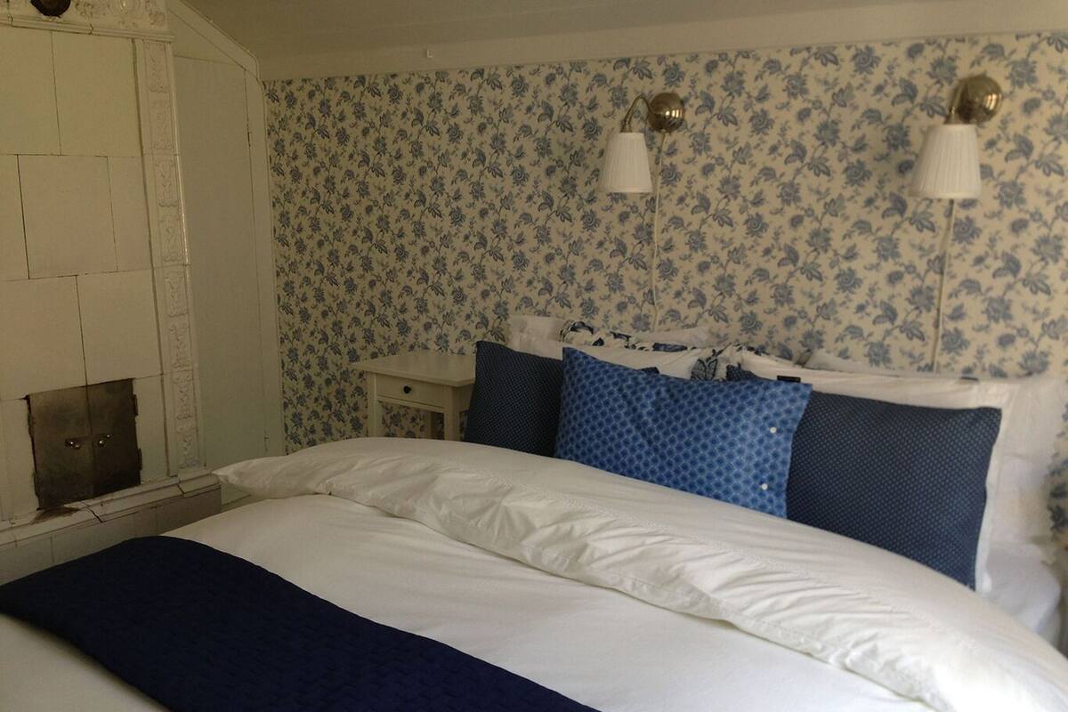 Una cama bien hecha con ropa de cama blanca y azul, una mesita de noche pequeña y una pared con estampado floral.
