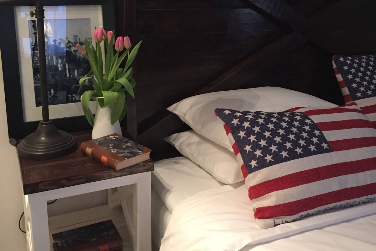Un dormitorio con cabecera de madera oscura, mesita de noche con lámpara, un cuadro enmarcado, un florero con flores y almohadas con diseño de la bandera estadounidense.
