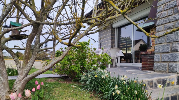Un edificio de piedra con un jardín delantero, que incluye un árbol, flores y un comedero para pájaros.