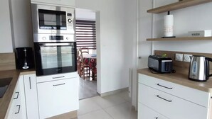 Cocina moderna con armarios blancos, horno integrado, microondas y cafetera. Se ve un área de comedor al fondo, con una mesa y sillas.