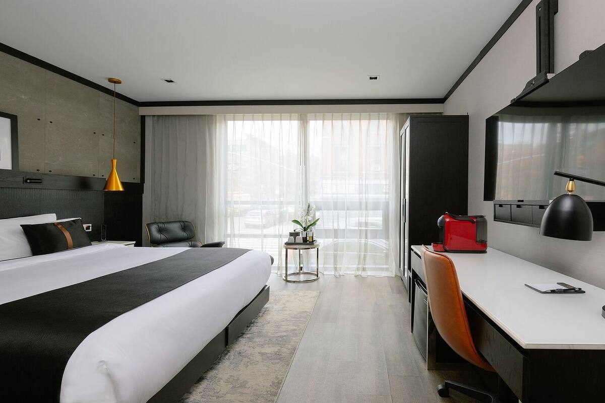 A modern hotel room with a bed, desk, chair, and a large window with sheer curtains.