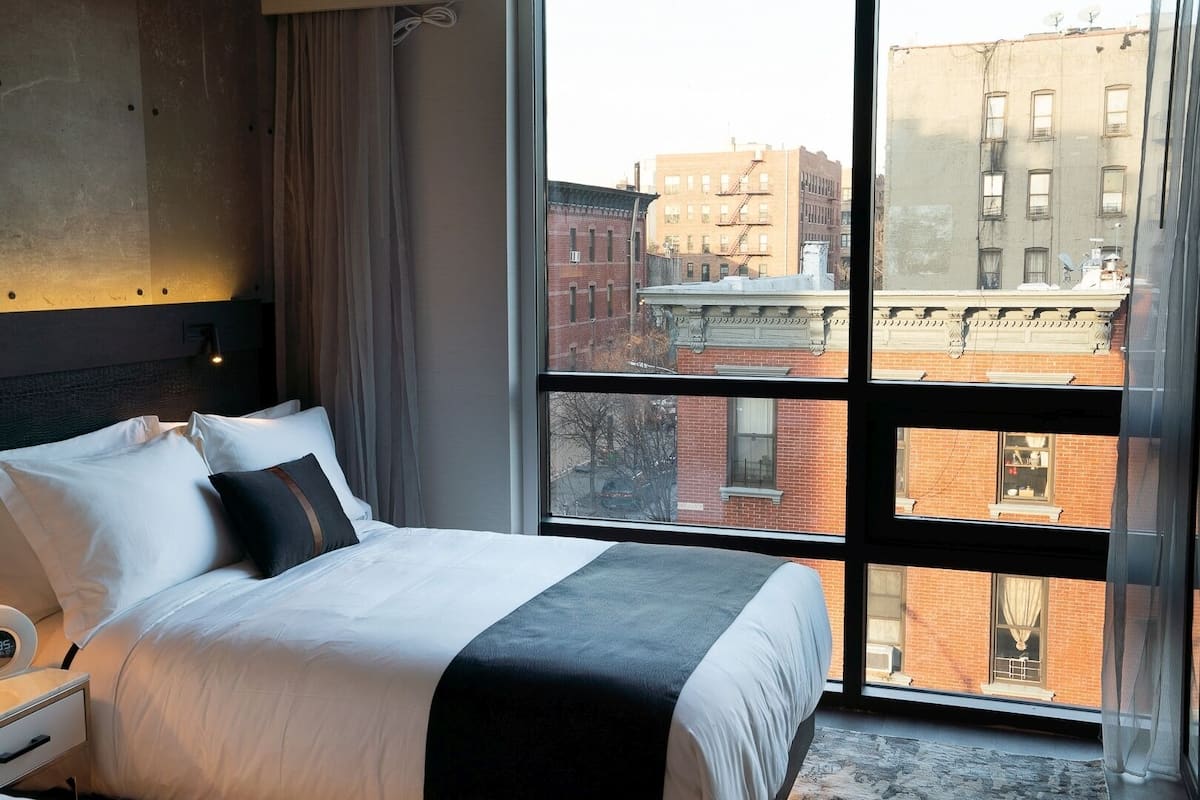 A hotel room with a large window, a bed with white bedding, a bedside table, and a wall-mounted light fixture.