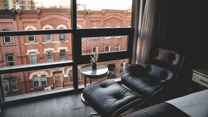 A room with a large window, a black leather chair, a small round table with a vase, and a view of red brick buildings. - 42 Hotel (Brooklyn)