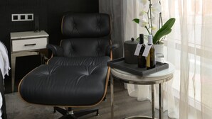 A modern room with a black leather chair, a small table with a bottle and a plant, a white bedside table with a remote control, and a window with sheer curtains. - 42 Hotel (Brooklyn)
