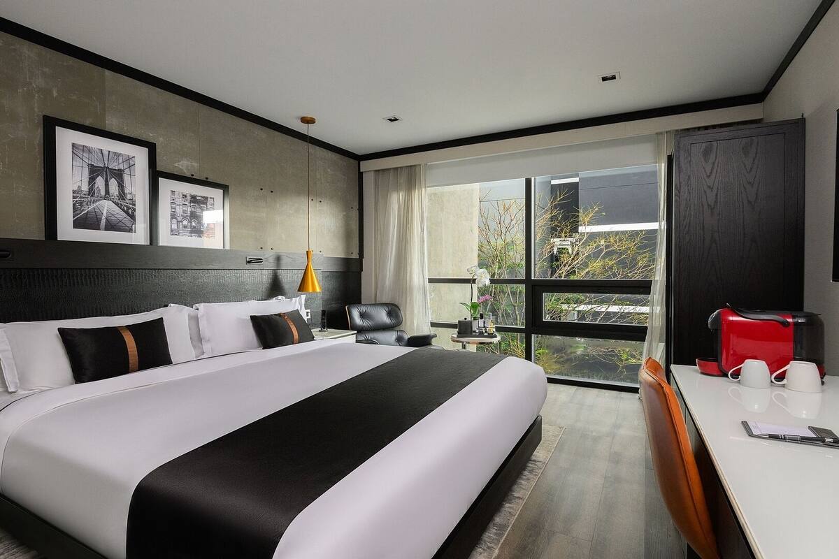 A modern hotel room with a large bed, a desk with a red appliance, and a view through a large window.