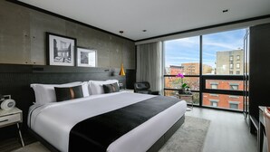 A modern hotel room with a large bed, a nightstand, a desk, a chair, a window with a city view, and two framed pictures on the wall. - 42 Hotel (Brooklyn)
