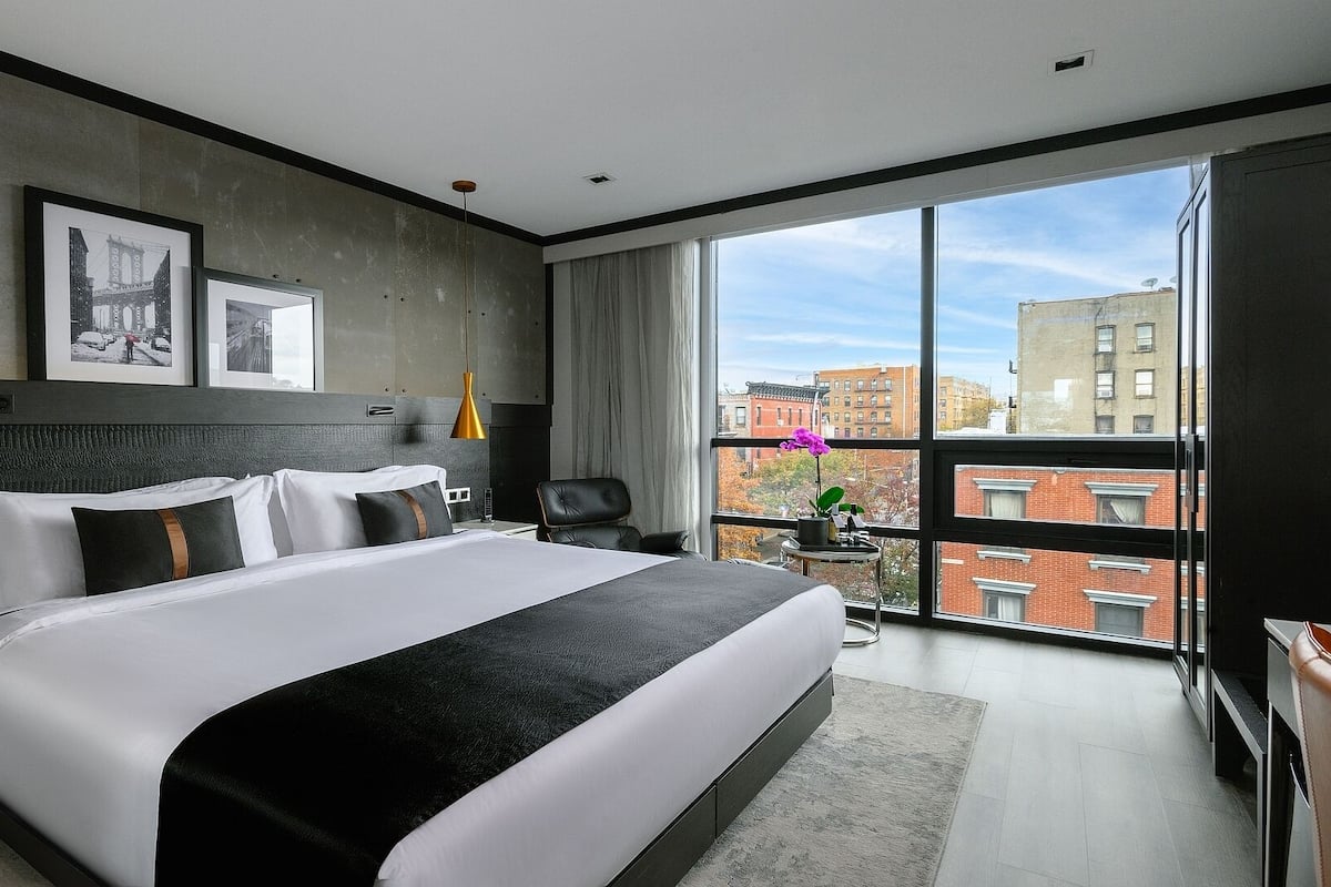 A modern hotel room with a large bed, a nightstand, a desk, a chair, a window with a city view, and two framed pictures on the wall.