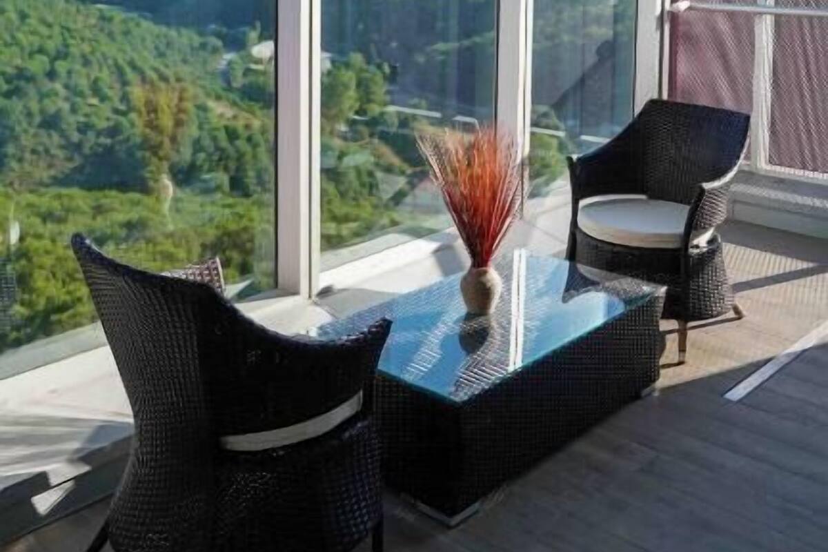 Un balcon avec une table en verre et deux fauteuils en osier, donnant sur une vallée boisée.