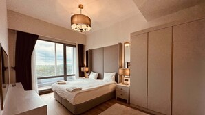 A modern hotel room with a large bed, a bedside table with lamps, a wardrobe, and a view of the city through large windows.